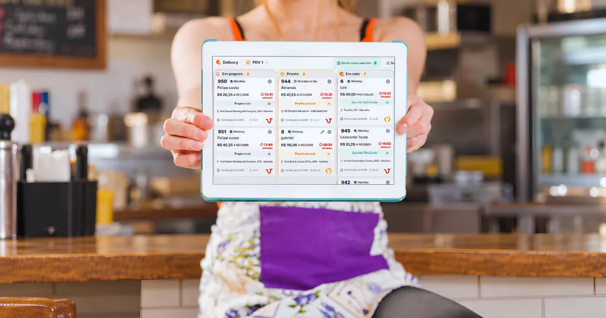 Mulher segura tablet com a Central de Delivery do ControleNaMão na tela, diante do balcão de um restaurante.