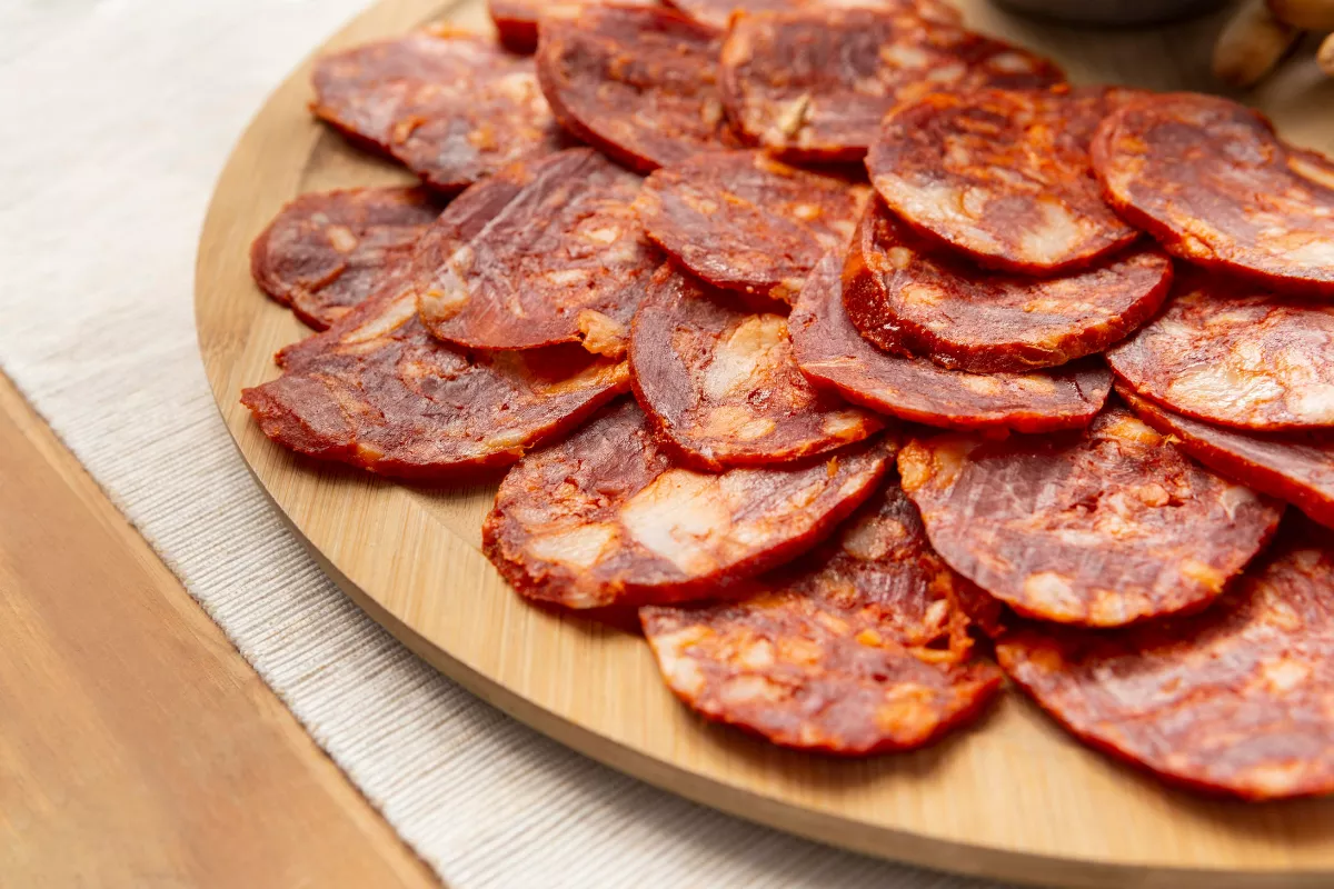 Fatias de calabresa uniformemente cortadas dispostas em uma tábua de madeira, representando a importância do cortador de frios para padronizar os ingredientes da pizza.