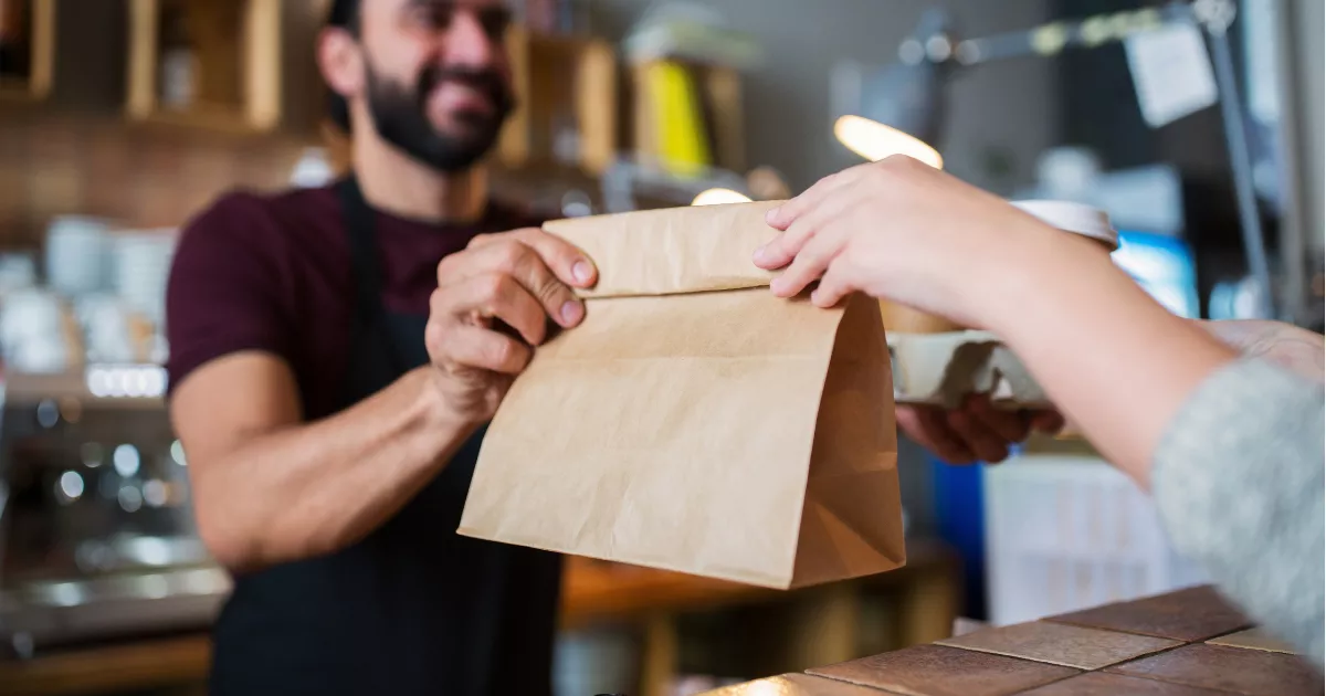 Atendente entrega uma sacola de papel a uma cliente no balcão de um restaurante, ilustrando a retirada de pedidos.