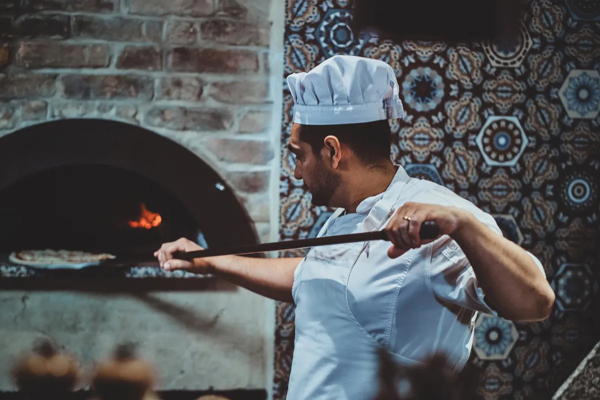 Pizzaiolo colocando pizza em forno a lenha tradicional, representando o cuidado no preparo e a importância de gestão e precificação justa em pizzarias.