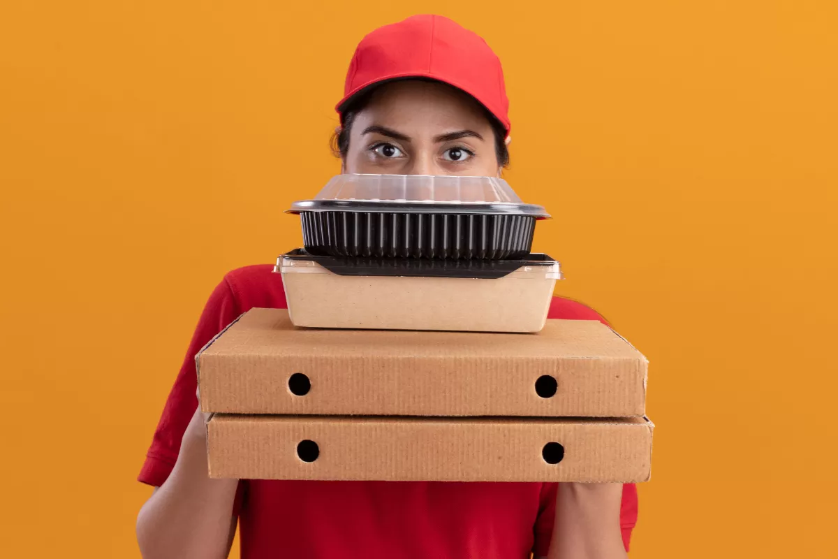 Uma entregadora de delivery, vestindo camisa e boné vermelhos, segura caixas de pizza e outros recipientes de delivery, ilustrando a importância das embalagens no transporte de alimentos.