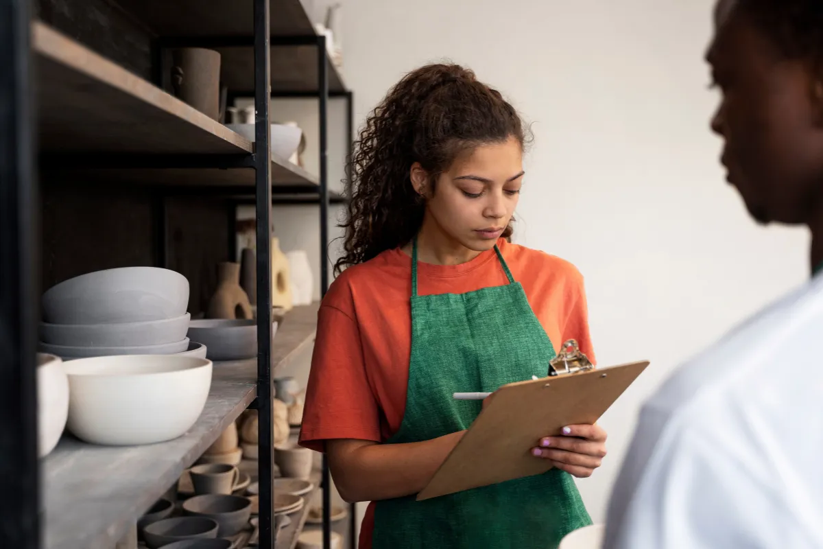 Uma mulher de avental verde e camiseta laranja anota informações em uma prancheta, conferindo o estoque de louças de cerâmica. O foco é em uma gestão de estoque estratégica para a Black Friday, garantindo que não faltem insumos no restaurante.