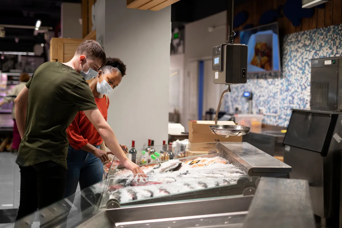 Compradores selecionando peixes frescos em supermercado, reforçando a importância da escolha e do recebimento correto de ingredientes para evitar perdas.