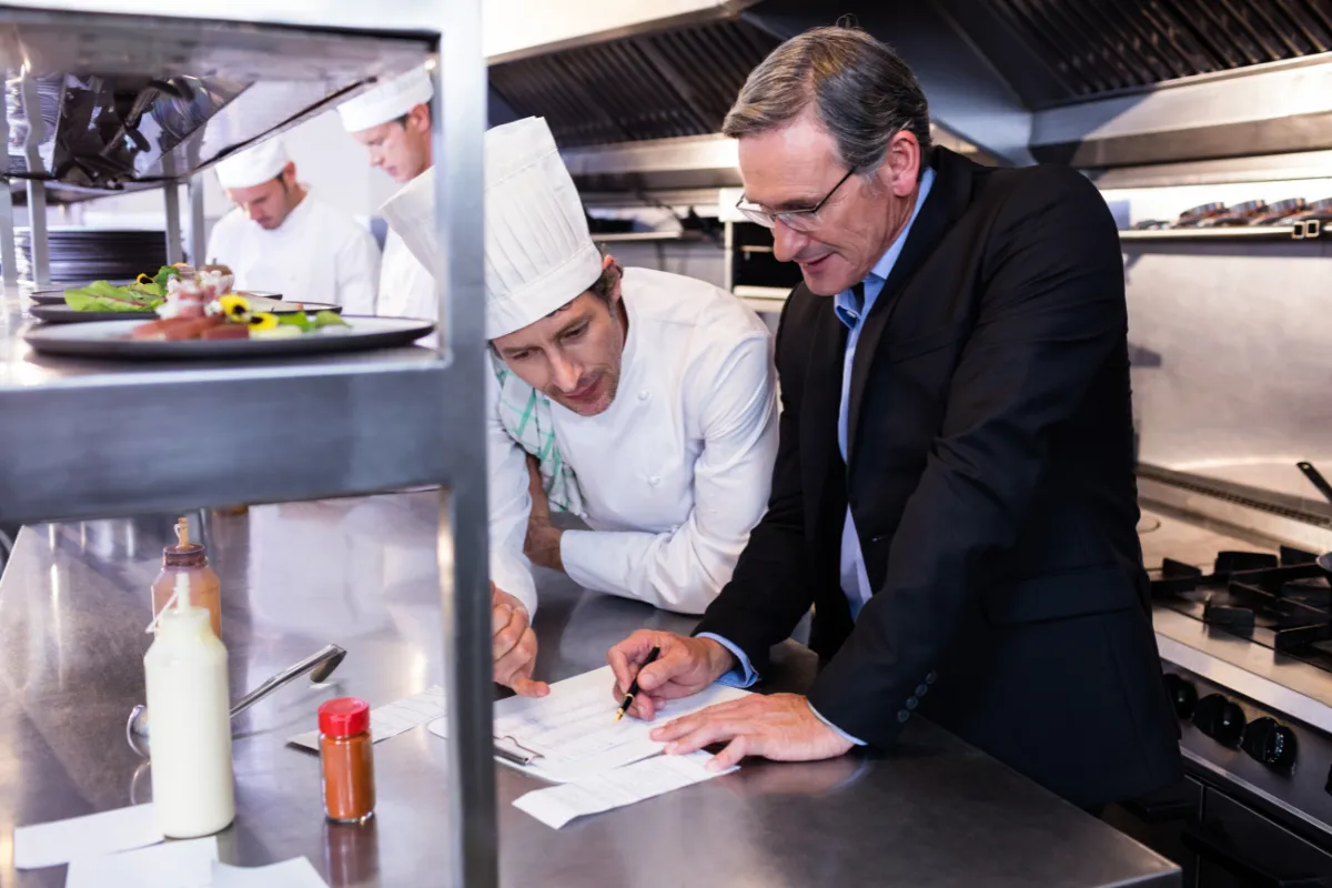 Gerente e chef analisando fichas técnicas na cozinha, representando a importância da gestão e do controle de produção para reduzir o desperdício de alimentos no restaurante.