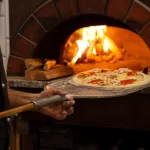 Um pizzaiolo coloca uma pizza fresca com cogumelos e pimentões em um forno a lenha aceso, destacando o uso do forno tradicional e a pá de pizza na cozinha.