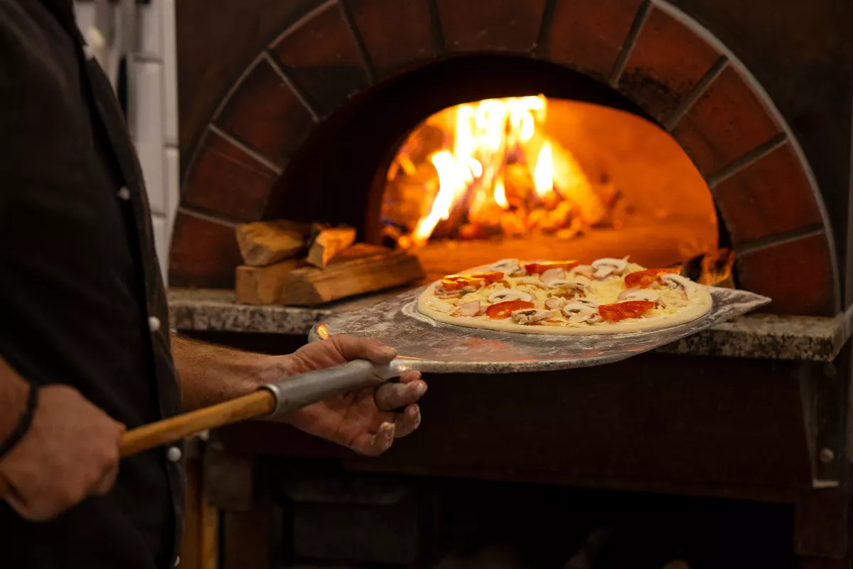 Um pizzaiolo coloca uma pizza fresca com cogumelos e pimentões em um forno a lenha aceso, destacando o uso do forno tradicional e a pá de pizza na cozinha.