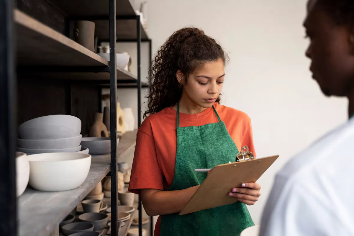 Uma mulher com avental verde anota informações em uma prancheta, em frente a prateleiras com utensílios, representando o controle de estoque e a gestão de insumos em uma pizzaria.