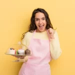 Jovem confeiteira com avental rosa comemora com um prato de doces na mão, ilustrando o sucesso ao entender e melhorar a margem de lucro da doceria.