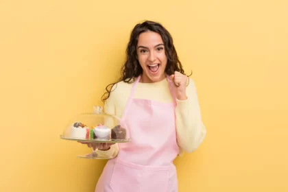 Jovem confeiteira com avental rosa comemora com um prato de doces na mão, ilustrando o sucesso ao entender e melhorar a margem de lucro da doceria.