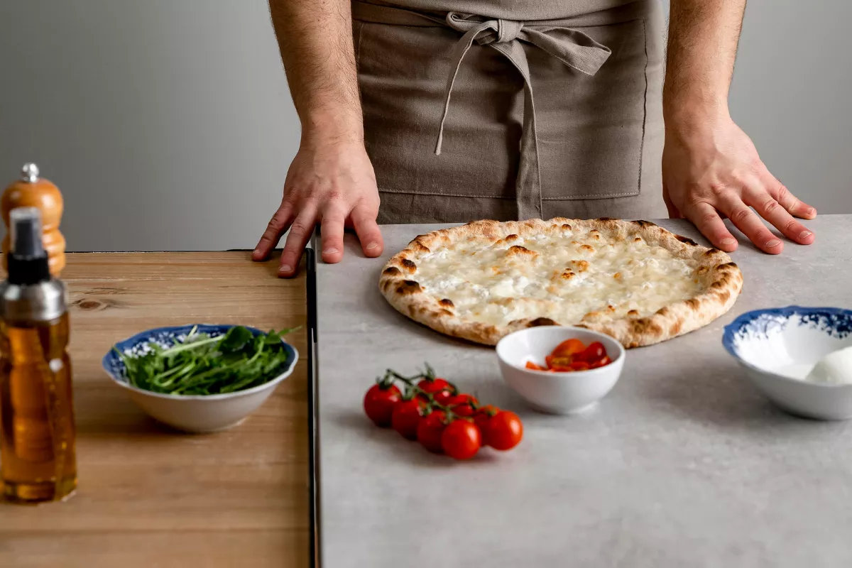 Mãos de um pizzaiolo em uma bancada de preparação, com uma pizza recém-assada e ingredientes frescos como tomates e rúcula, ilustrando a montagem e finalização das pizzas.