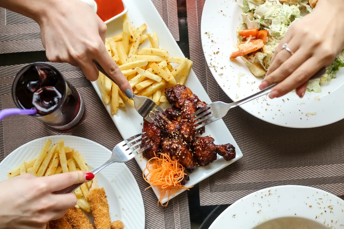 Duas pessoas, com as mãos visíveis, compartilham uma porção de asinhas de frango com batatas fritas no centro de uma mesa. A imagem representa o conceito de "combos" e ofertas promocionais para atrair clientes durante a Black Friday.