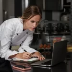 Chef de restaurante consultando informações no computador e anotando dados fiscais, exemplificando a importância do controle financeiro na escolha da alíquota do Simples Nacional.