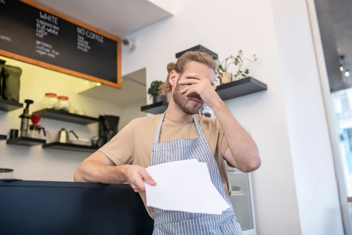 Um homem, com uniforme de barista e avental, esconde o rosto com uma das mãos, parecendo sobrecarregado. A imagem representa o desafio de gerir a equipe e o estresse do time durante picos de movimento, como o da Black Friday.