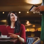 Uma mulher sorridente em um restaurante, vestindo um vestido vermelho e segurando um cardápio da mesma cor, enquanto um garçom, de costas, anota o pedido em uma comanda. A cena sugere o momento de escolha ou pedido no estabelecimento.
