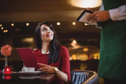 Uma mulher sorridente em um restaurante, vestindo um vestido vermelho e segurando um cardápio da mesma cor, enquanto um garçom, de costas, anota o pedido em uma comanda. A cena sugere o momento de escolha ou pedido no estabelecimento.