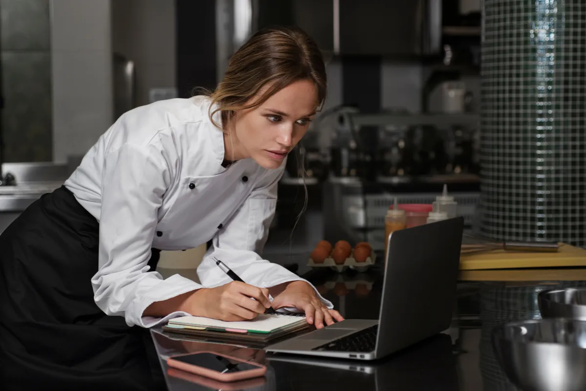 Uma mulher com uniforme de chef, em uma cozinha profissional, olha atentamente para a tela de um laptop, enquanto segura uma caneta e anota em um caderno. A imagem simboliza a fase de planejamento e estratégia para a Black Friday no restaurante.