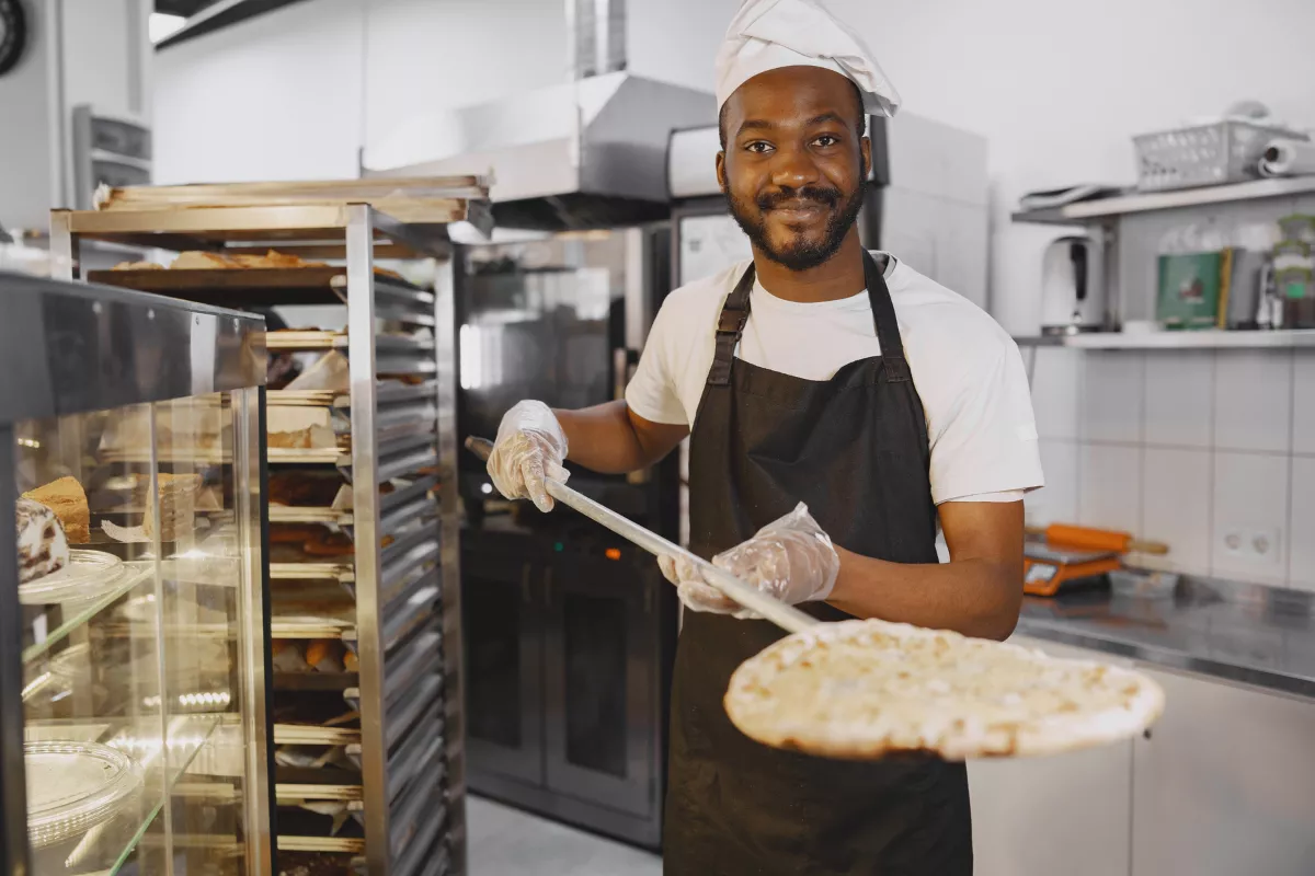 Um pizzaiolo sorridente, usando um chapéu de chef e avental, segura uma pizza crua na pá, pronto para levá-la ao forno, mostrando a etapa de preparo e cozimento.
