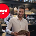 Empresário de restaurante sorrindo com cardápio em mãos, ao lado de um notebook, com o logo do iFood em destaque — contexto sobre investir no iFood ADS.