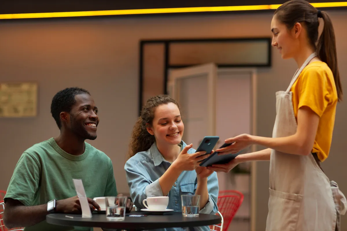 Dois clientes sorrindo em um restaurante, com a mulher pagando a conta usando um celular via Pix, enquanto a garçonete, também sorridente, segura uma maquininha ou tablet para o pagamento. O homem ao lado observa a transação.
