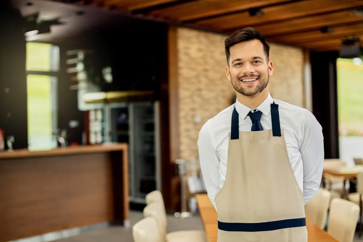 Garçom sorridente de avental em um salão de restaurante, representando o papel de atendimento ao cliente na operação do restaurante.