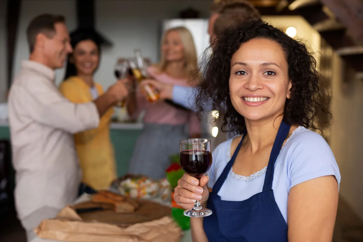 Mulher com avental segurando uma taça de vinho enquanto clientes brindam ao fundo, simbolizando a interação entre equipe e experiência do cliente.