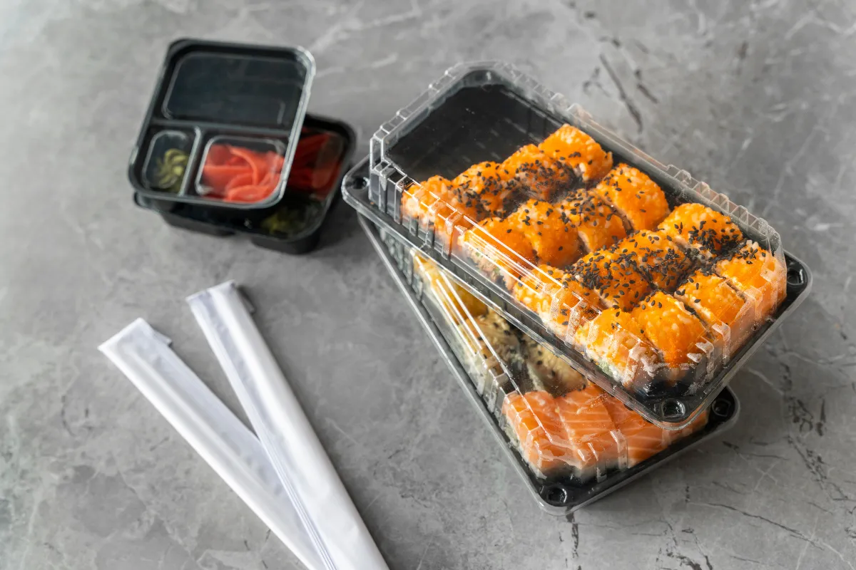 Embalagens de plástico transparente com sushi e molhos sobre uma mesa cinza, representando o uso do polipropileno (PP) e do PET em deliveries de comida japonesa e refeições frias, com boa vedação e praticidade.