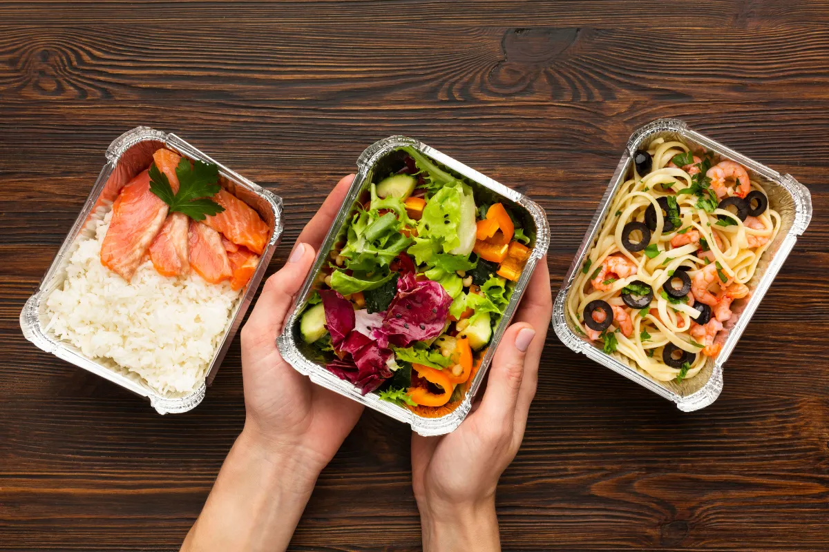 Três marmitas de alumínio abertas com salmão, salada e macarrão sobre uma mesa de madeira, ilustrando embalagens térmicas usadas em deliveries de pratos quentes como grelhados, massas e refeições executivas.