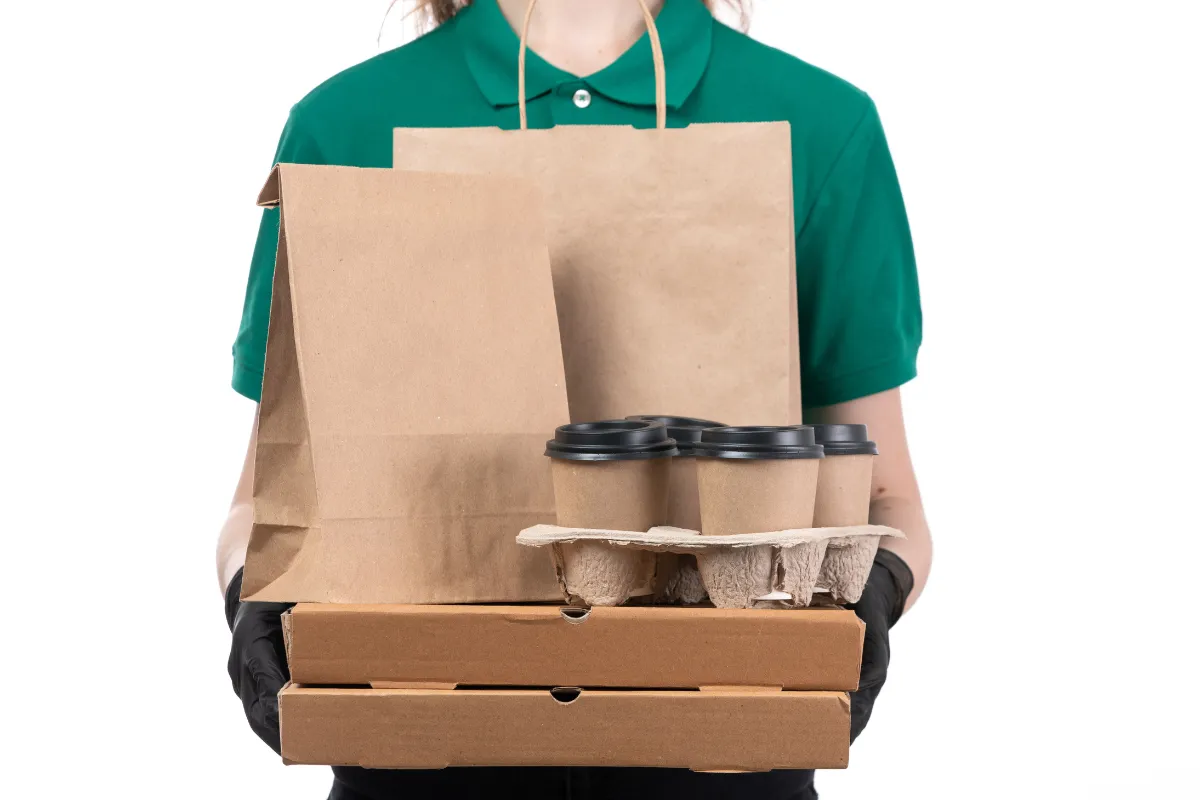 Entregador com camisa verde segurando caixas de pizza, copos de café e sacolas de papelão, destacando o uso do papelão como material térmico e resistente em deliveries de pizzarias, cafeterias e fast foods.