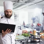 Chef de cozinha usando tablet enquanto prepara um prato, representando o uso de monitor KDS para organizar pedidos e otimizar o fluxo de trabalho no restaurante.