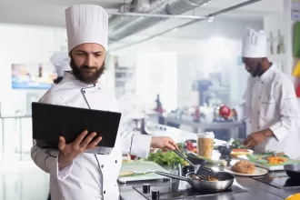 Chef de cozinha usando tablet enquanto prepara um prato, representando o uso de monitor KDS para organizar pedidos e otimizar o fluxo de trabalho no restaurante.