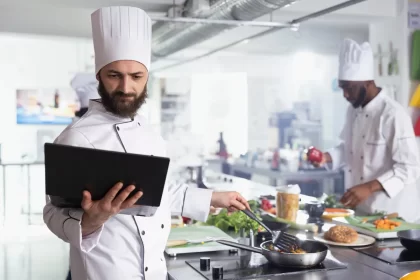 Chef de cozinha usando tablet enquanto prepara um prato, representando o uso de monitor KDS para organizar pedidos e otimizar o fluxo de trabalho no restaurante.