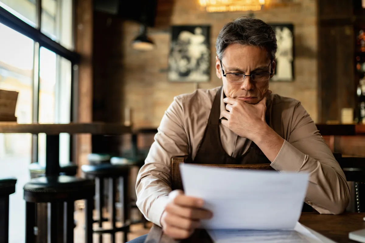 Dono de restaurante com expressão séria analisando um papel, representando a preocupação e o estudo sobre o novo Imposto sobre Valor Agregado (IVA).