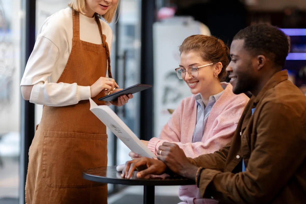 Garçonete usa tablet para anotar pedido de casal em restaurante, simbolizando a modernização do atendimento e a experiência otimizada na jornada do cliente.