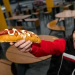 Cliente satisfeito segurando hot dog com salsicha e maionese em lanchonete, demonstrando experiência positiva no delivery