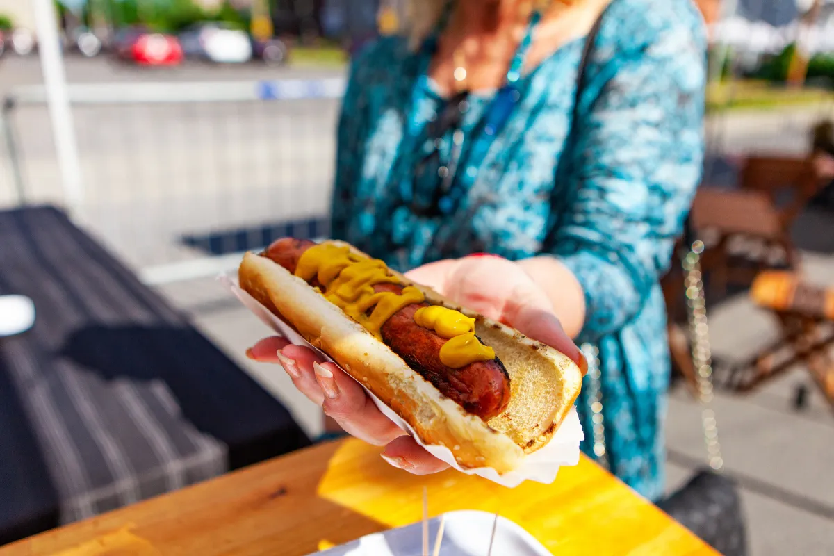 Pessoa segurando hot dog simples com mostarda em embalagem descartável, ideal para delivery de cachorro-quente
