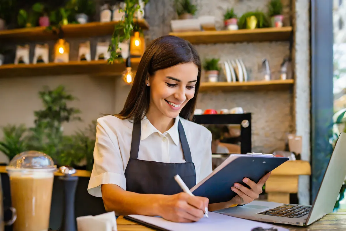 Mulher, gerente ou proprietária de restaurante, sorri enquanto preenche uma prancheta, revisando o planejamento financeiro e o cronograma de adaptação do negócio à nova Reforma Tributária.