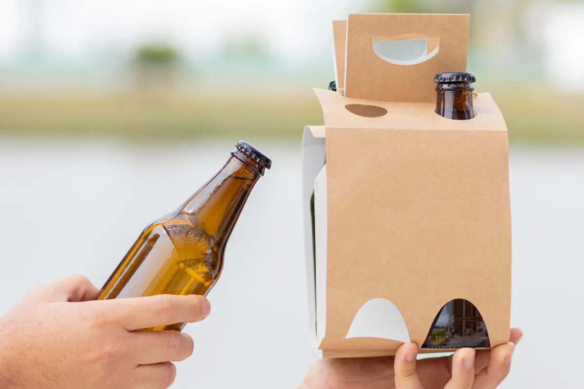 Duas pessoas trocando garrafas de cerveja, uma delas segurando um suporte de papel kraft com divisórias, mostrando embalagens seguras para transporte de bebidas no delivery e prevenção de choques e vazamentos.