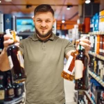 Dono de bar satisfeito em um corredor de bebidas, segurando duas garrafas, o que simboliza a importância da logística de entrega e pontualidade de uma distribuidora para a gestão de estoque.