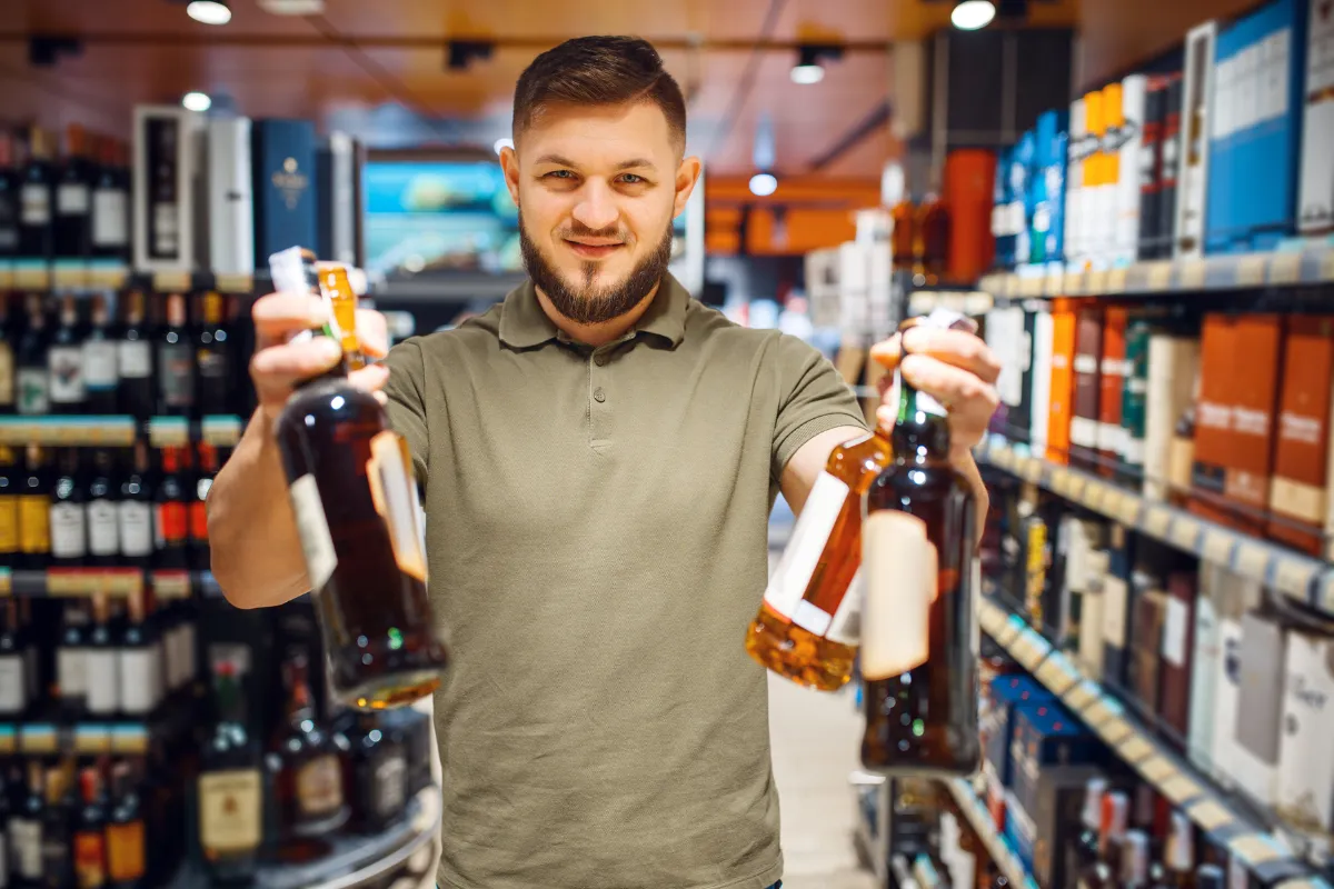 Dono de bar satisfeito em um corredor de bebidas, segurando duas garrafas, o que simboliza a importância da logística de entrega e pontualidade de uma distribuidora para a gestão de estoque.