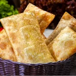 Cesto de vime escuro transbordando com pastéis fritos e crocantes, com legumes frescos e pimentas vermelhas suavemente desfocados ao fundo, reforçando a ideia de comida fresca e saborosa.