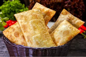 Cesto de vime escuro transbordando com pastéis fritos e crocantes, com legumes frescos e pimentas vermelhas suavemente desfocados ao fundo, reforçando a ideia de comida fresca e saborosa.