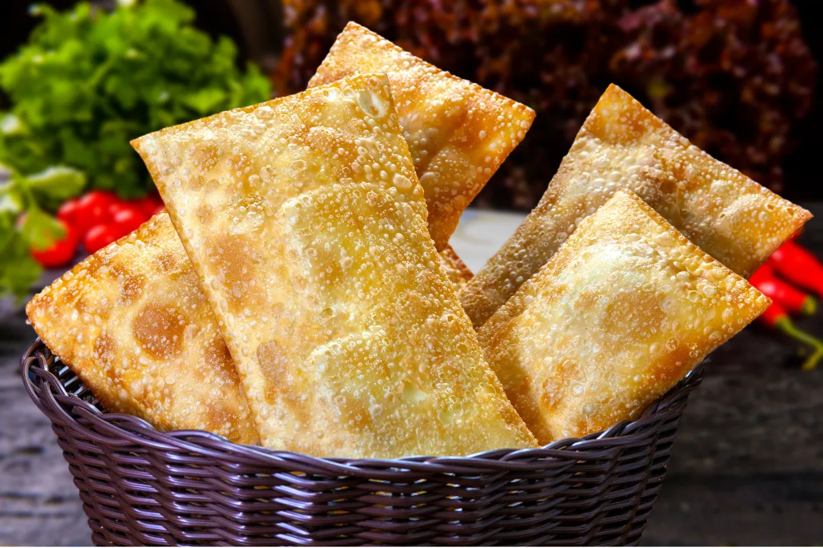 Cesto de vime escuro transbordando com pastéis fritos e crocantes, com legumes frescos e pimentas vermelhas suavemente desfocados ao fundo, reforçando a ideia de comida fresca e saborosa.