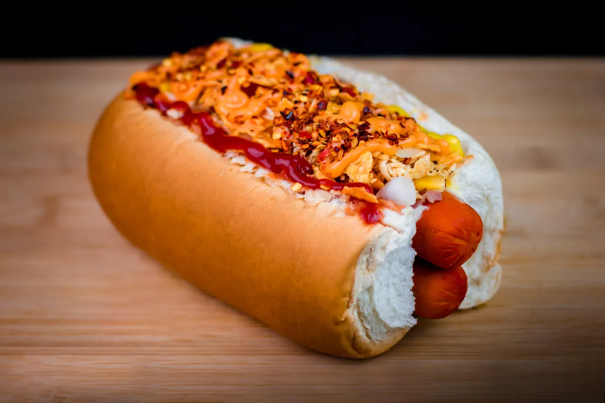 Hot dog completo com salsicha, molho de tomate, maionese temperada e temperos sobre pão macio em superfície de madeira