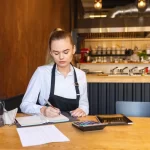 Jovem funcionária de restaurante, vestindo avental e camisa social, sentada à mesa com documentos e calculadora, dedicando-se à tarefa de entender e planejar as mudanças trazidas pela Reforma Tributária de 2026.