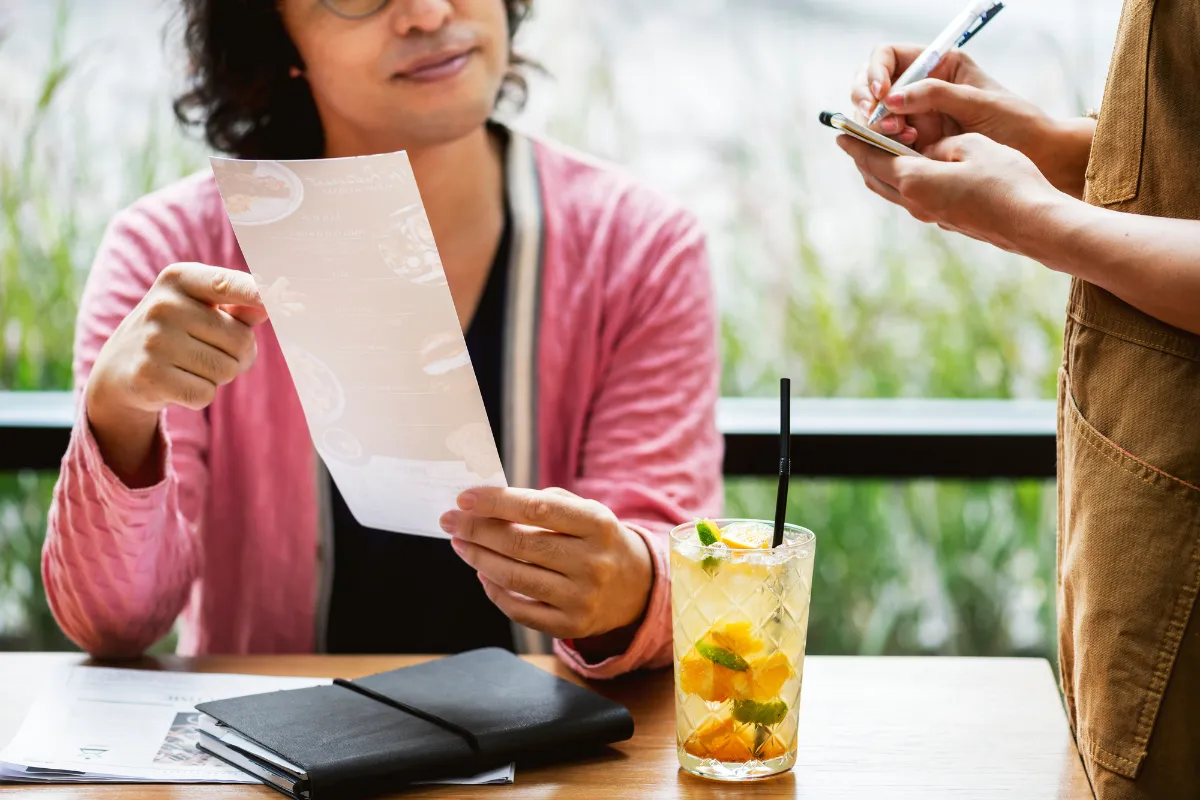 Cliente consultando um cardápio enquanto um garçom anota o pedido ao lado de uma bebida. Representa o serviço e a aplicação das novas alíquotas do IVA em bares e restaurantes.