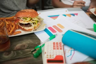 Tabela de relatórios, gráficos de barras coloridos e uma calculadora ao lado de um hambúrguer gourmet e batatas fritas, simbolizando a análise do Percentual de Crescimento do faturamento do restaurante, um dos principais indicadores de desempenho.