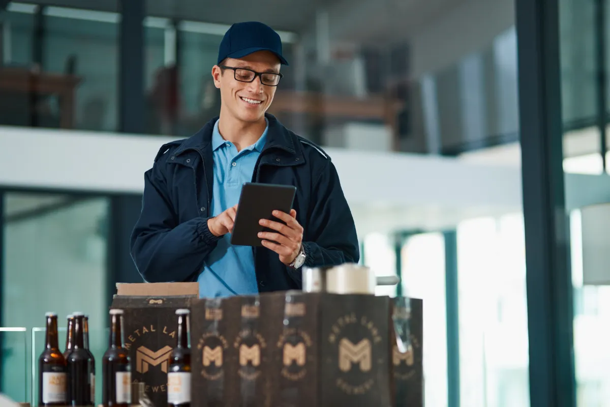 Entregador de uma distribuidora de bebidas sorrindo enquanto usa um tablet para conferir o pedido de um bar, ilustrando a importância de avaliar o custo-benefício, preço e condições ao escolher um fornecedor.