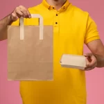 Pessoa vestida com camisa amarela segurando uma sacola e uma caixa de papel kraft sobre fundo rosa, representando embalagens sustentáveis e personalizáveis para lanches e deliverys ecológicos.
