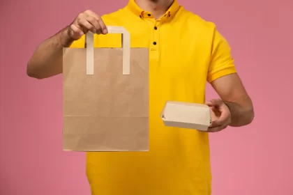 Pessoa vestida com camisa amarela segurando uma sacola e uma caixa de papel kraft sobre fundo rosa, representando embalagens sustentáveis e personalizáveis para lanches e deliverys ecológicos.