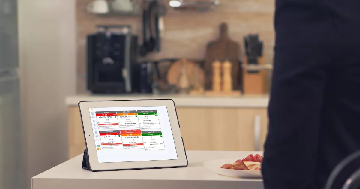 Tablet apoiado em bancada de cozinha exibindo o monitor KDS do ControleNaMão com pedidos organizados por mesa e status de preparo em vermelho e verde